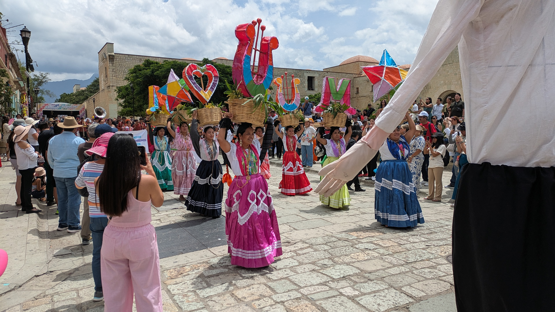 A colorful street in Oaxaca City during a festival, with dancers in traditional attire under a clear blue sky, surrounded by colonial architecture and vibrant markets.