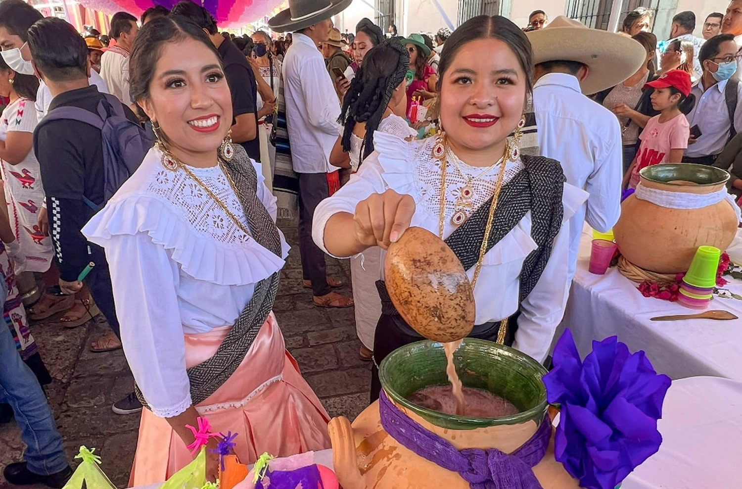 Young women in traditional Oaxacan dress offering cups of agua fresca from a flower-adorned stand during Día de la Samaritana celebrations