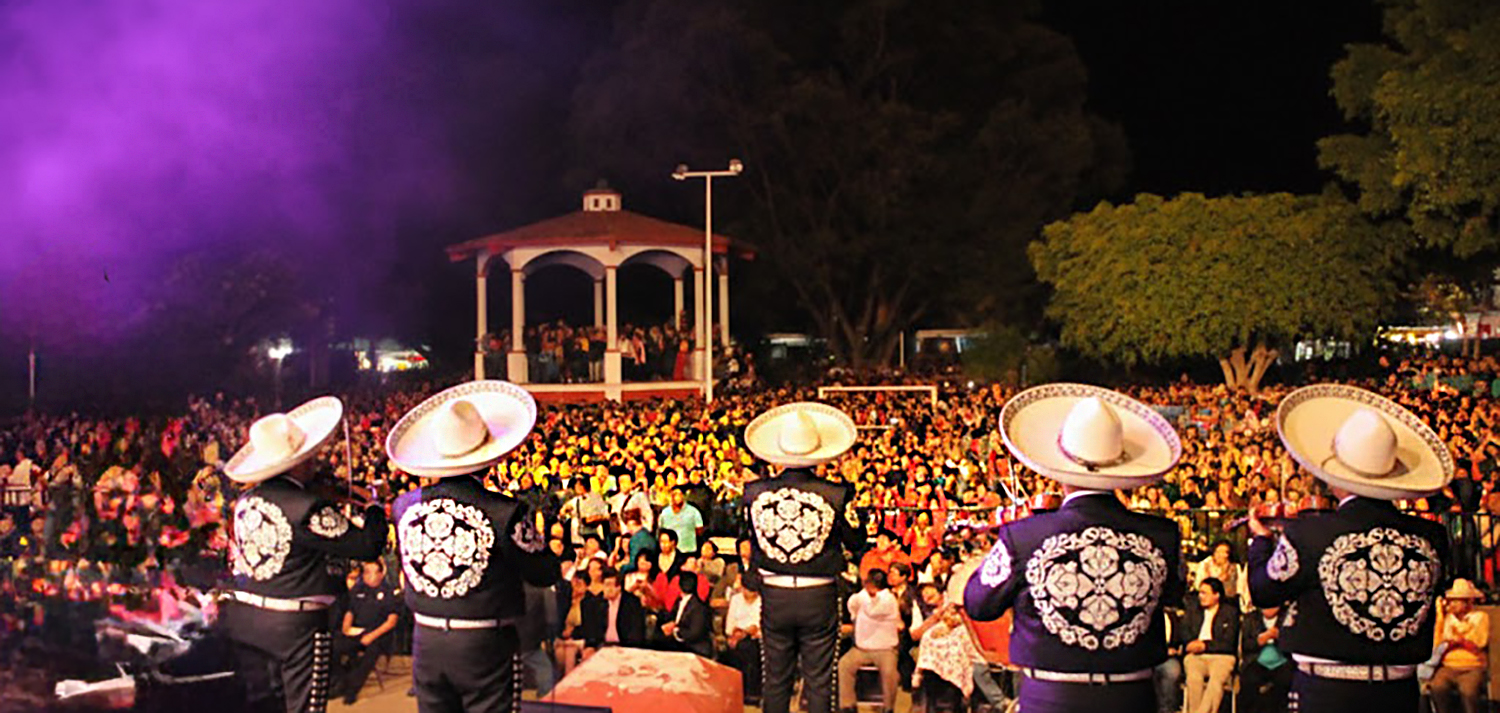 Nighttime crowd gathered in the main plaza of Santa Cruz Xoxocotlán during Martes de Brujas with a stage and lights visible
