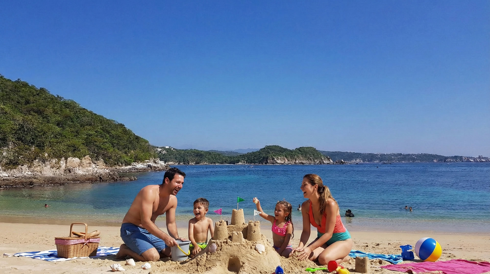 A family building sandcastles on a serene bay beach in Huatulco, Oaxaca, with calm turquoise waters and lush green hills in the background